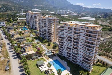 Exteriorul proprietății din Alanya prezintă o vilă galbenă cu turn, cu accente de piatră, un gazon luxuriant și o potecă care duce spre mare.