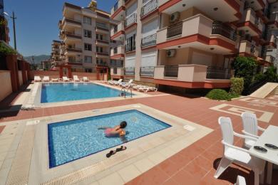 Балкон у нерухомасці ў Аланіі з плеценай мэбляй, шкляным сталом, кафлянай падлогай і каванай жалезнай агароджай, які прапануе светлае жыццё на адкрытым паветры.