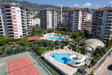 Alanya eiendom balkongutsikt med utsikt over bassenger, hager og bybygninger under en lys, solrik himmel.