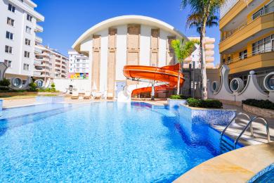 Alanya'da turkuaz denize ve güneşli sahil şeridine bakan hasır mobilyalı ve cam korkuluklu balkon.