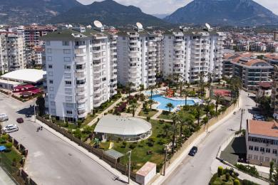 ملک آلانیا: اتاق نشیمن روشن و با پلان باز با مبل بژ، کف کاشی کاری شده و آشپزخانه سفید مدرن.