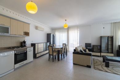 Zona de zi luminoasă, de tip open-space, a apartamentului din Alanya are o bucătărie modernă, un set de dining și scaune confortabile sub o lumină caldă.