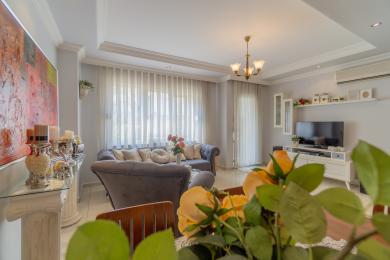 Proprietatea din Alanya are o sufragerie cu o canapea gri de pluș, perdele transparente și o atmosferă luminoasă și aerisită.