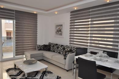 Alanja nekretnina dnevna soba sa modernim kaučem, trpezarijom i elegantnim horizontalnim žaluzinama, stvarajući svetlu i prozračnu atmosferu.