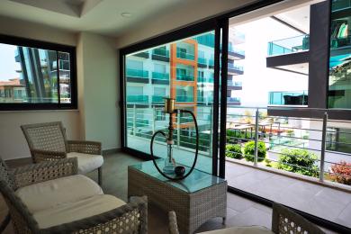 Bright Alanya balcony with wicker furniture, glass table, and sliding doors overlooking modern buildings.