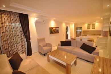 Alanya property living room featuring neutral sofas, tile flooring, and an open-plan kitchen with warm, inviting lighting.