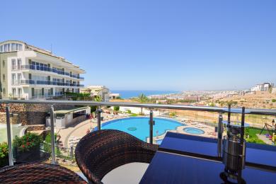 Alanya ejendom altan med kurvemøbler med udsigt over en pool, by og Middelhavet på en solskinsdag.
