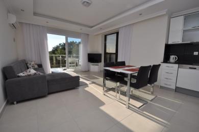 Heller Wohnbereich in einem Apartment in Alanya mit grauem Sofa, Esstisch und moderner weißer Küche unter natürlichem Licht.
