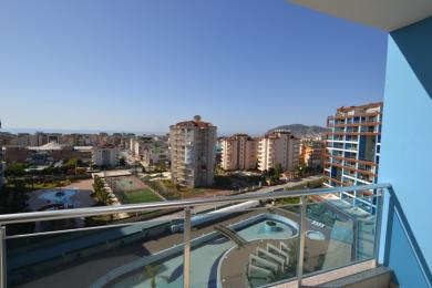 Alanya Immobilien Balkonblick mit Blick auf Stadt, Pool und Meer unter hellem Sonnenlicht, der eine entspannte Atmosphäre schafft.