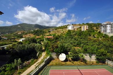 Alanya eiendomsvisning som viser en tennisbane med fjell- og bygningsutsikt under en blå himmel.