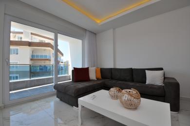 Obývací pokoj v Alanyi s mramorovou podlahou, plyšovou šedou pohovkou a přístupem na balkon, vytvářející světlou a moderní atmosféru.