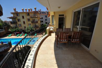 Balkon nekretnine u Alanji sa podom od travertina, ogradom od kovanog željeza i pogledom na bazen, namješten drvenim trpezarijskim setom.