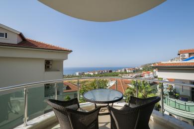 Alanya eign með svölum með húsgögnum úr wicker og glerhandriði, sem býður upp á töfrandi útsýni yfir hafið og borgina í björtu sólarljósi.