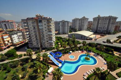 Alanya'da satılık daire manzarası: Palmiye ağaçları ve apartman binalarıyla çevrili, kaydıraklı, tatil köyü tarzı havuz, güneşli bir atmosfer.