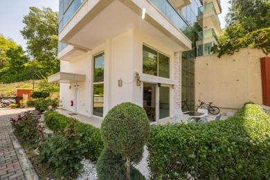 Außenansicht einer Immobilie in Alanya mit einer modernen weißen Fassade, großen Fenstern und einem angelegten Garten mit Sitzgelegenheiten im Freien.