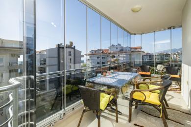 Cam duvarları, hasır mobilyaları ve şehir manzarası ile aydınlık Alanya balkonu, rahatlatıcı ve güneşli bir atmosfer yaratıyor.