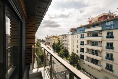 Balkon nemovitosti v Alanyi se skleněným zábradlím, dřevěnými prvky a výhledem na město pod zataženou oblohou.