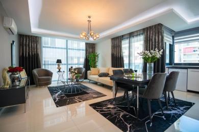 Alanya-Immobilienwohnzimmer mit einem modernen Kronleuchter, Marmorteppichen und raumhohen Fenstern mit transparenten Vorhängen.