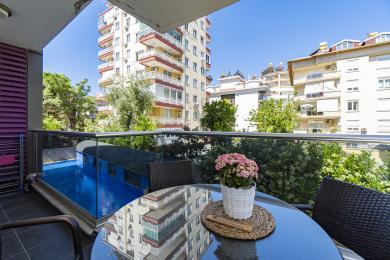 Alanya eiendom balkong med glassrekkverk, kurvstoler og utsikt over bassenget, badet i sterkt sollys.