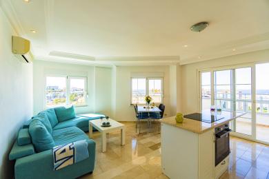 Világos alanyai apartman nappali türkizkék kanapéval, márványpadlóval és tengerre néző erkéllyel.