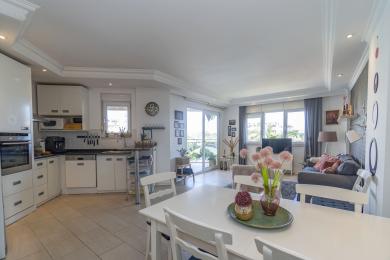 Aydınlık, açık planlı Alanya dairesi, modern beyaz mutfak, yemek alanı ve balkon erişimi olan oturma odasına sahiptir.