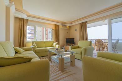 Alanya Immobilie Wohnzimmer mit bequemen Sitzgelegenheiten, einem Glastisch, Zugang zum Balkon und warmem, einladendem Licht.