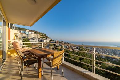 Alanya eign með svölum með viðarborði og stólum sem bjóða upp á töfrandi útsýni yfir hafið og borgina í björtu og sólríku andrúmslofti.