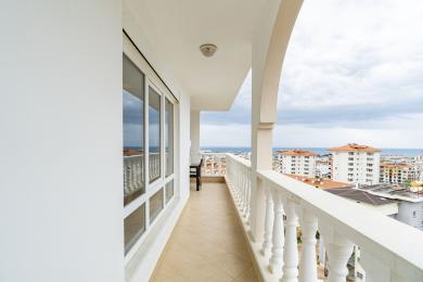Alanya-Immobilienbalkon mit weißer Balustrade, Fliesenboden und Stadtblick unter einem bewölkten Himmel.