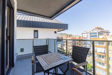Alanya संपत्ति में विकर फर्नीचर, टाइल टेबल, ग्लास रेलिंग और उज्ज्वल, धूप वाले शहर के दृश्यों वाला बालकनी।