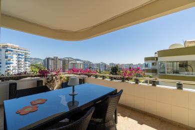 Alanya balcony with wicker furniture, glass table, and vibrant bougainvillea overlooking the city skyline.
