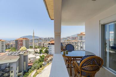 Balkon nekretnine u Alanji sa nameštajem od ratana i staklenim stolom, nudi svetao pogled na grad i more.