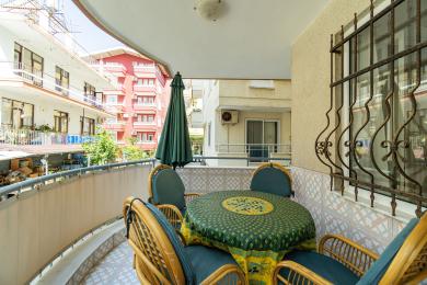 Alanya eiendom balkong med rottingmøbler, mønstret duk og sterkt dagslys, som skaper en koselig utendørs spiseplass.