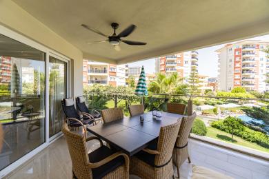 Balkon w Alanyi z meblami wiklinowymi, wentylatorem sufitowym i jasnymi, słonecznymi widokami na basen i ogrody.