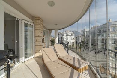 Balkon nekretnine u Alanji sa staklenim zidovima, podom od pločica, ležaljkama i pogledom na grad, stvarajući svijetlu i opuštajuću atmosferu.