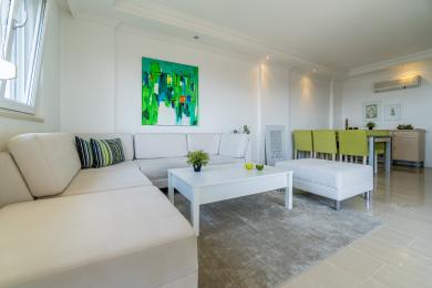 Svetlá obývacia izba v Alanyi s bielou pohovkou, moderným umením a jedálenským kútom, vytvárajúca čistú a vzdušnú atmosféru.