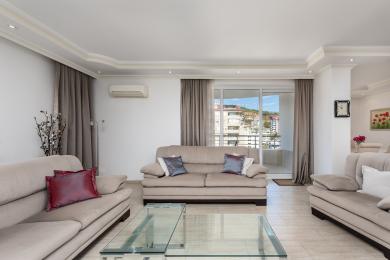 Helles Wohnzimmer in Alanya mit beigen Sofas, Glastisch, Zugang zum Balkon und neutralen Vorhängen, die eine gemütliche Atmosphäre schaffen.