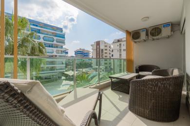 Alanya ejendom balkon med kurvemøbler, glasrækværk og lyse, solrige udsigter over nærliggende bygninger.