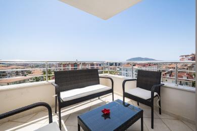 Proprietate în Alanya cu balcon mobilat cu mobilier din răchită, perne albe și vedere panoramică asupra orașului sub lumina puternică a soarelui.
