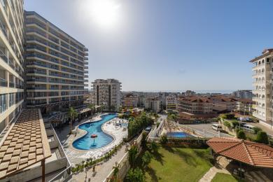 Alanya eiendomsvisning: Balkong med utsikt over et stort basseng, byens skyline og Middelhavet på en solrik dag.