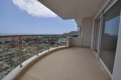 Prostrani balkon u Alanji s podovima od pločica, staklenim ogradama i panoramskim pogledom na grad i more, okupan sunčevom svjetlošću.