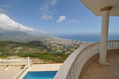 Balkón nehnuteľnosti v Alanyi s kamenným zábradlím, bazénom a panoramatickým výhľadom na pobrežie pod jasnou oblohou.