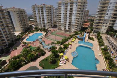 Proprietatea din Alanya, cu vedere de la balcon, prezintă piscine, terenuri de tenis și peisaje luxuriante sub lumina puternică a soarelui, creând o atmosferă de stațiune.