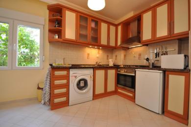 صورة لمطبخ في عقار في ألانيا يتميز بخزائن خشبية دافئة، لمسات كريمية، باكسبلاش من البلاط، وإضاءة طبيعية تخلق جواً مريحاً.